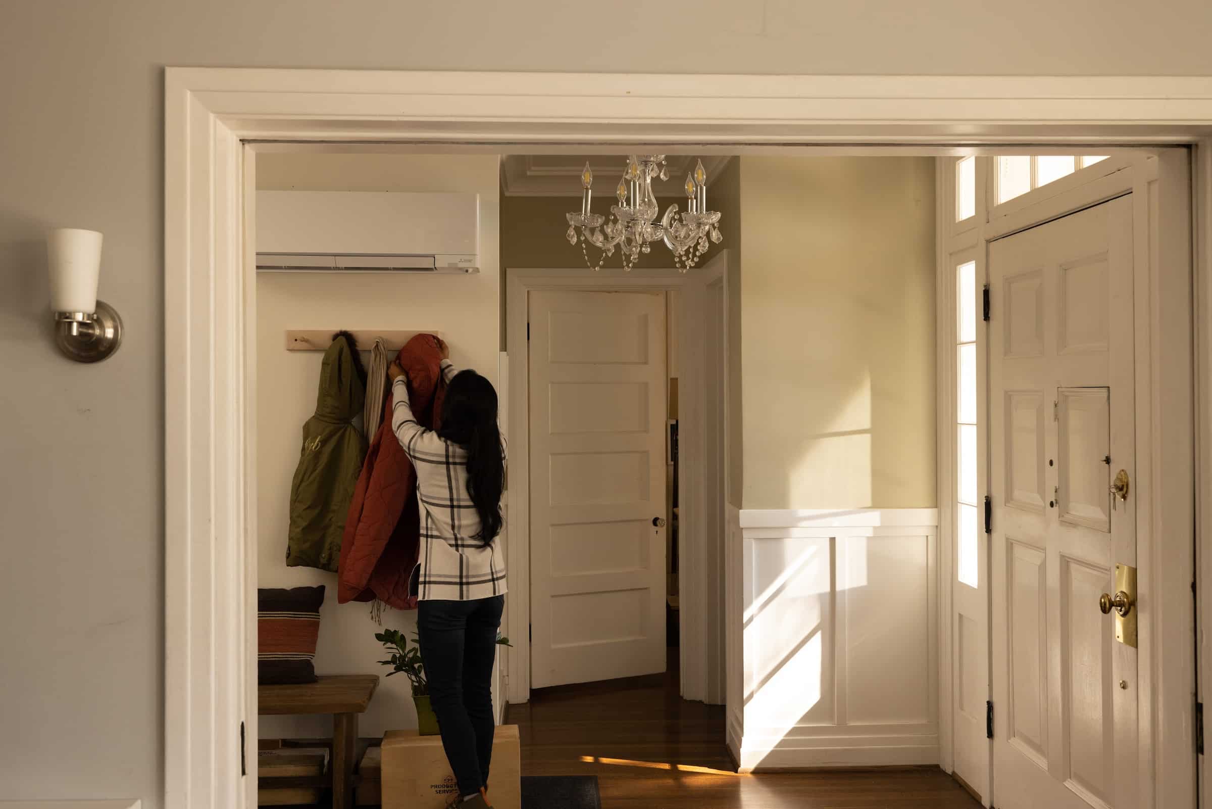 Woman hanging up coat on a coat rack near the door. A Mitsubishi wall mount heat pump is installed on the wall above her