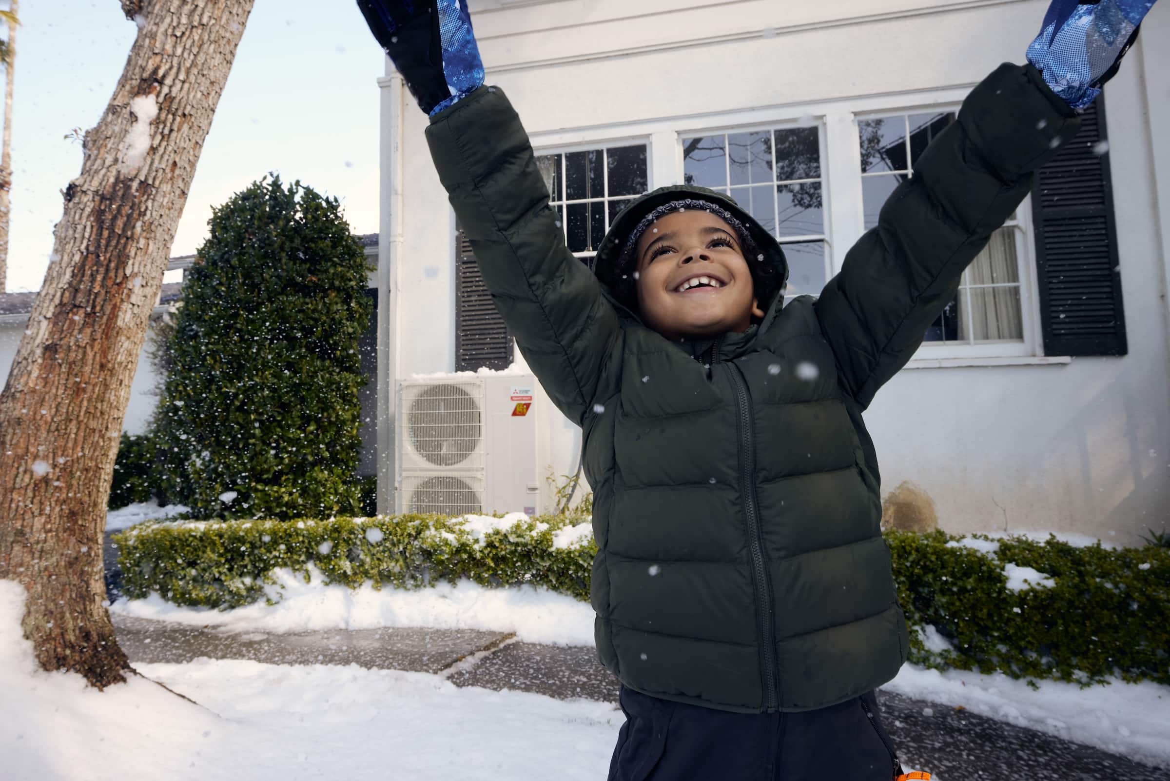 Child playing outside a house in the snow. A Mitsubishi heat pump is sitting outside of the house in the background.