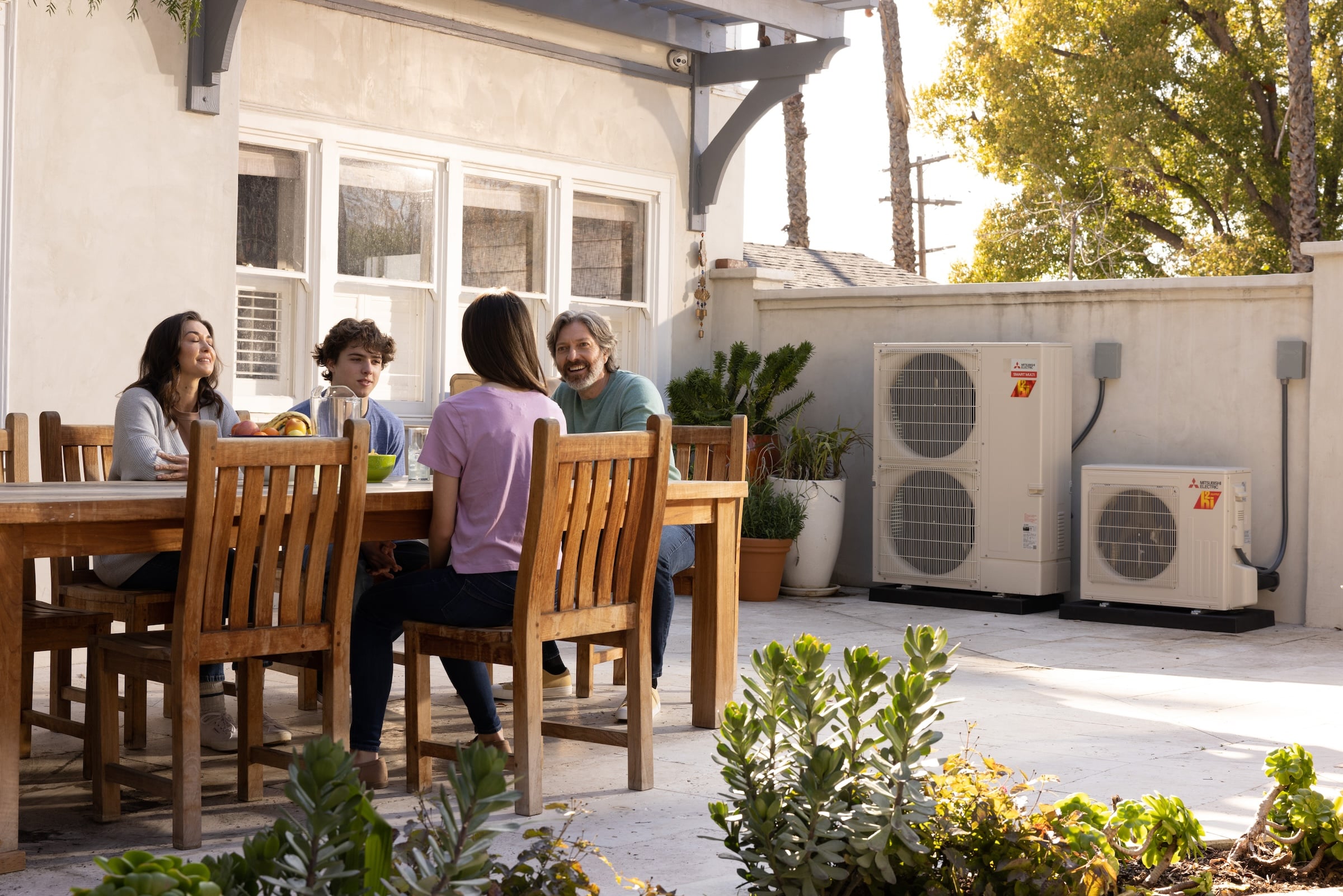 Family sitting at table outside in back yard with Mitsubishi heat pumps setup outside of the house in the background